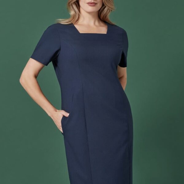 A person stands confidently wearing a navy blue, short-sleeved dress with a square neckline. Their hand is in a pocket, and they pose against a solid green background.