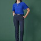 A person stands against a green background, wearing a blue blouse and dark trousers. They have hands in their pockets and are looking slightly upwards. The outfit is completed with black shoes and a belt.