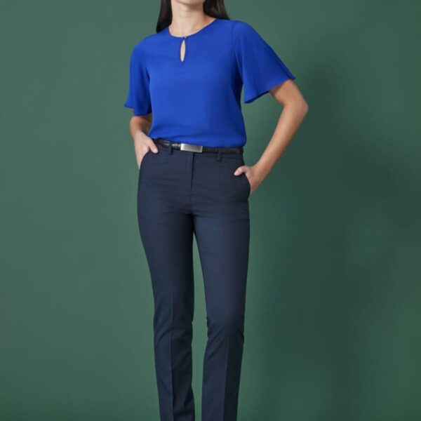 A person stands against a green background, wearing a blue blouse and dark trousers. They have hands in their pockets and are looking slightly upwards. The outfit is completed with black shoes and a belt.