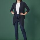 A woman stands against a dark green background, wearing a navy cardigan, white blouse, navy pants, and black heels. Her long dark hair is worn down, and she has a confident expression with her hands by her sides.