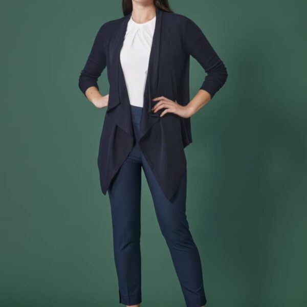 A woman stands against a dark green background, wearing a navy cardigan, white blouse, navy pants, and black heels. Her long dark hair is worn down, and she has a confident expression with her hands by her sides.