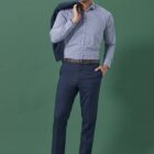 A man in a blue checkered shirt and dark blue trousers, holding a blue jacket over his shoulder, stands against a green background. He is wearing brown shoes and a brown belt.