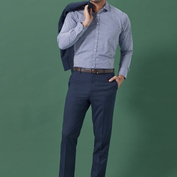A man in a blue checkered shirt and dark blue trousers, holding a blue jacket over his shoulder, stands against a green background. He is wearing brown shoes and a brown belt.