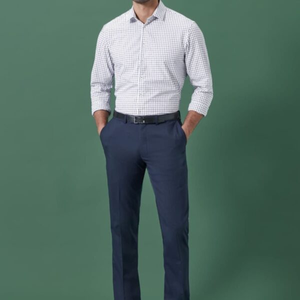 A man stands against a green background, wearing a checkered shirt, dark blue trousers, and black shoes. His hands are in his pockets, and he is facing the camera.