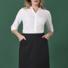A person with long hair stands against a green background, wearing a white button-up shirt with three-quarter sleeves tucked into a knee-length black skirt. They have hands in the skirt pockets and are looking confidently at the camera.