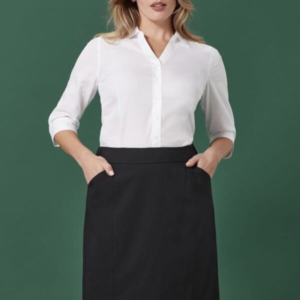 A person with long hair stands against a green background, wearing a white button-up shirt with three-quarter sleeves tucked into a knee-length black skirt. They have hands in the skirt pockets and are looking confidently at the camera.
