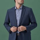 A person wearing a navy blue suit and a light blue striped shirt stands against a green background, buttoning the blazer while looking to the side.