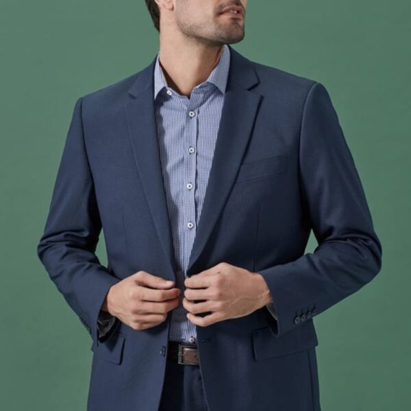 A person wearing a navy blue suit and a light blue striped shirt stands against a green background, buttoning the blazer while looking to the side.
