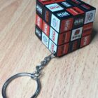 A Rubiks cube keychain sits on a wooden surface. The cube features various red, black, and white logos, including SUS, NORDECA, and TOYOTA, on its stickers. The keychains metal ring is attached to one corner of the cube.