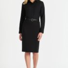 A person wearing a black long-sleeve dress with a collar and belt stands against a plain white background. They are wearing black shoes and have long, wavy hair.