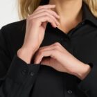 A person wearing a black button-up shirt is posing with their hands near their face. The focus is on the shirts buttons and the persons hands. Their hair is light and slightly wavy.