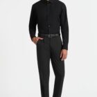A person standing against a white background, wearing a black long-sleeve shirt and black trousers. The individual has one hand in their pocket and is wearing black shoes and a belt.