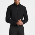 Smiling person wearing a black dress shirt and black pants, standing against a plain white background. Their hands are clasped together in front of them.