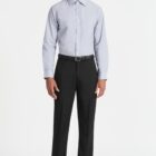 A person wearing a light blue dress shirt, black trousers, and black shoes is standing and smiling against a plain white background.