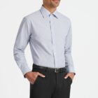 A person stands in a light blue button-up shirt, with hands in the pockets of their black pants. They have short hair and a mustache, wearing a black belt. The background is plain white.