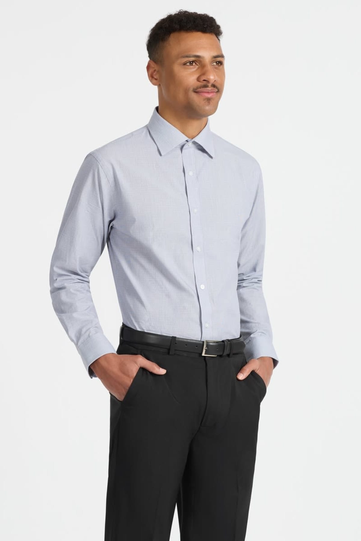 A person stands in a light blue button-up shirt, with hands in the pockets of their black pants. They have short hair and a mustache, wearing a black belt. The background is plain white.