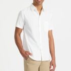 A person wearing a white short-sleeve button-up shirt and beige shorts stands against a plain white background, smiling with one hand in their pocket.