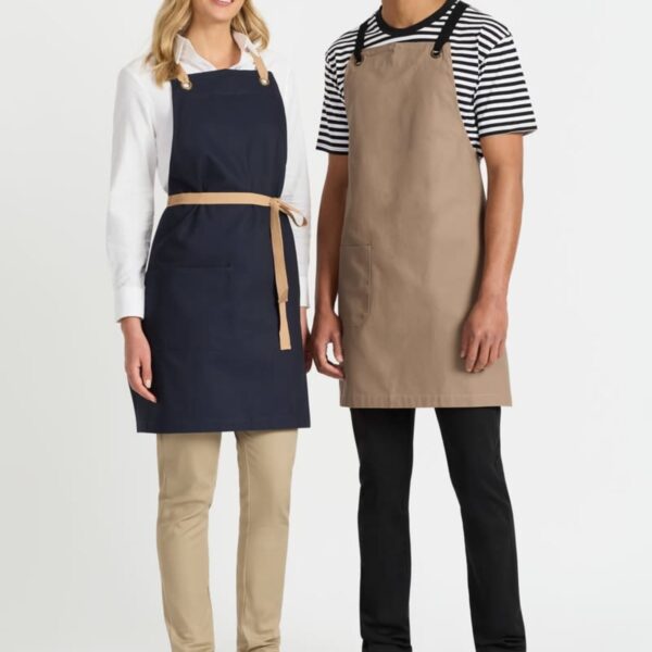 A woman and a man wearing aprons stand side by side, smiling. The woman has a black apron with a waist tie, while the man wears a brown apron. She has a white shirt and beige pants; he has a striped shirt and black pants. Both wear white shoes.