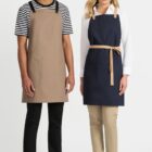 Two people stand side by side wearing aprons. The person on the left wears a tan apron over a striped shirt and black pants. The person on the right wears a navy apron over a white shirt and beige pants. Both wear white sneakers.