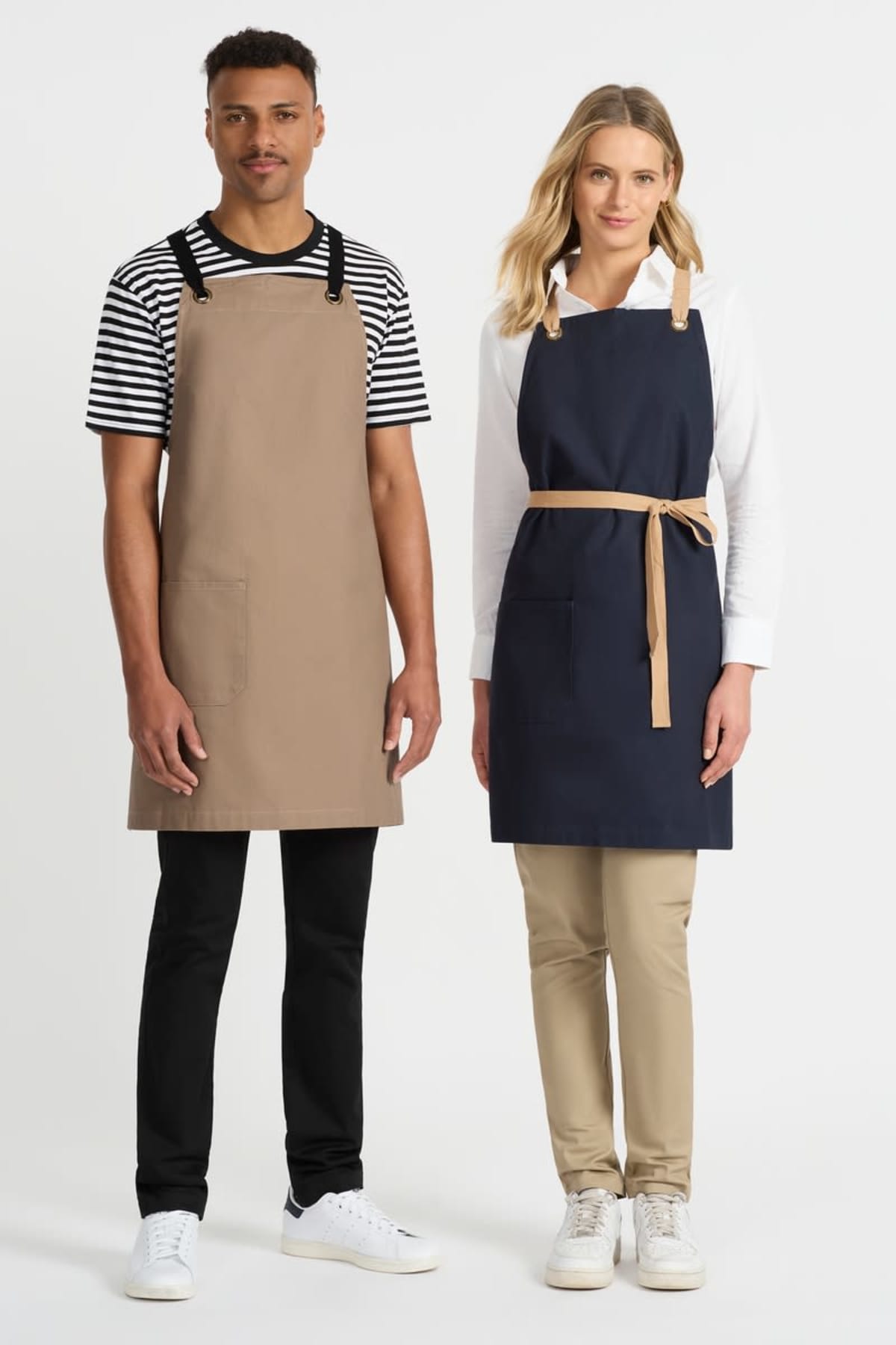 Two people stand side by side wearing aprons. The person on the left wears a tan apron over a striped shirt and black pants. The person on the right wears a navy apron over a white shirt and beige pants. Both wear white sneakers.