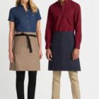 A woman and a man stand side by side, both smiling. The woman wears a denim short-sleeve shirt, a beige apron, black pants, and white sneakers. The man wears a red and black checkered shirt, a black apron, beige pants, and white sneakers.