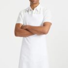 A person wearing a white short-sleeve shirt and a white apron stands confidently with arms crossed. The background is plain white, highlighting their welcoming smile.