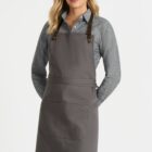 A person wearing a gray button-up shirt and a gray apron with dark brown straps. Their hands are behind their back, and they are standing against a plain white background.