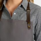 A person with blonde hair wears a gray button-up shirt and a gray apron with brown leather straps. The image shows a close-up view from the shoulders to the chest, capturing the upper part of the outfit.