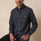 A person with short hair and a mustache is sitting on a stool against a beige background. They are wearing a dark button-up shirt and dark pants, looking directly at the camera with a slight smile.