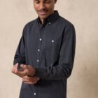 A person wearing a dark button-up shirt stands against a beige background. They are adjusting their sleeve with a gentle smile, looking down. The shirt has a pocket and white buttons.