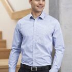 A person with short dark hair is wearing a light blue, long-sleeved button-up shirt with a subtle check pattern, paired with black pants. They are standing indoors near a staircase and looking to the side, smiling.