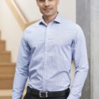 Man in a light blue, checkered dress shirt and black pants stands indoors near a staircase. He is smiling and has dark hair. The background features a light-colored wall and wooden steps.