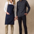 Two people stand side by side against a neutral backdrop. The person on the left wears a white shirt, navy apron, and beige pants. The person on the right wears a black button-up shirt and black pants. Both wear white sneakers and smile at the camera.