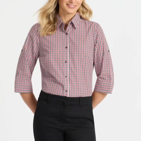 A person with long blonde hair is smiling, wearing a red and white checkered shirt with rolled-up sleeves and black pants. They are standing against a plain white background with their hands behind their back.