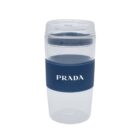 A clear travel mug with a blue silicone sleeve featuring the brand name PRADA in white. The lid is translucent, and the overall design is sleek and modern.