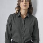 A person with long, wavy hair wearing a dark button-up shirt and black pants stands against a light gray wall, looking towards the camera. Their sleeves are rolled up to the elbows.