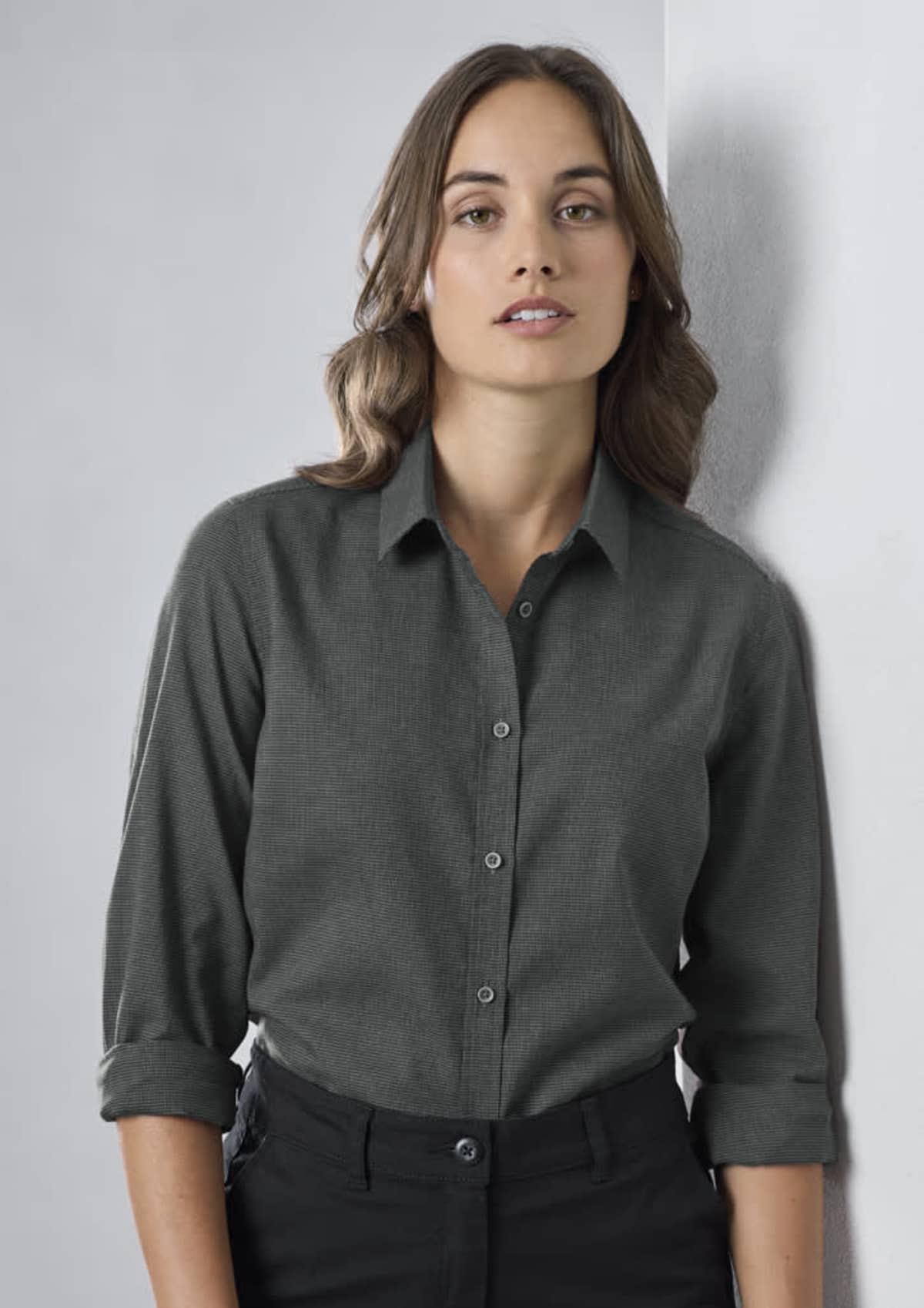 A person with long, wavy hair wearing a dark button-up shirt and black pants stands against a light gray wall, looking towards the camera. Their sleeves are rolled up to the elbows.