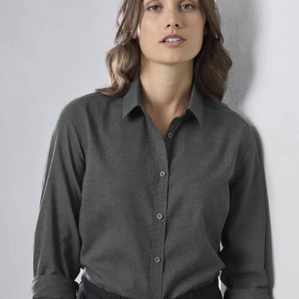 A person with long, brown hair wearing a gray button-up shirt with the sleeves rolled up. They are standing against a plain, light-colored background, looking directly at the camera with a neutral expression.