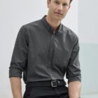 A man with short hair is sitting and wearing a gray button-up shirt with rolled-up sleeves and black pants. He is leaning slightly forward, with a neutral expression, in a softly lit setting.