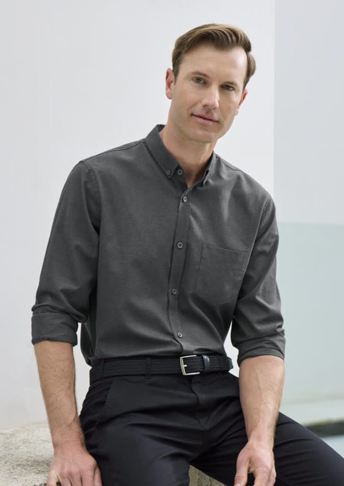 A man with short hair is sitting and wearing a gray button-up shirt with rolled-up sleeves and black pants. He is leaning slightly forward, with a neutral expression, in a softly lit setting.