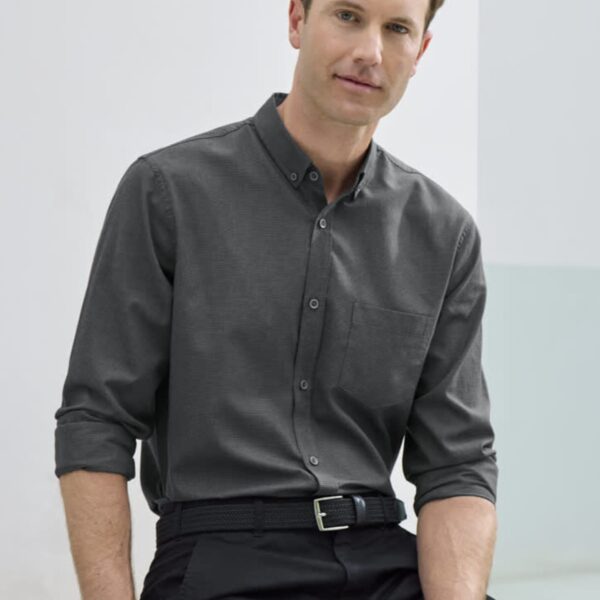 A man with short hair is wearing a dark button-up shirt and black pants, sitting casually against a light-colored wall. His sleeves are rolled up, and he has a relaxed expression.