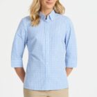 A person wearing a light blue and white checkered button-up shirt with three-quarter sleeves and beige pants stands against a plain white background, smiling with hands behind their back.