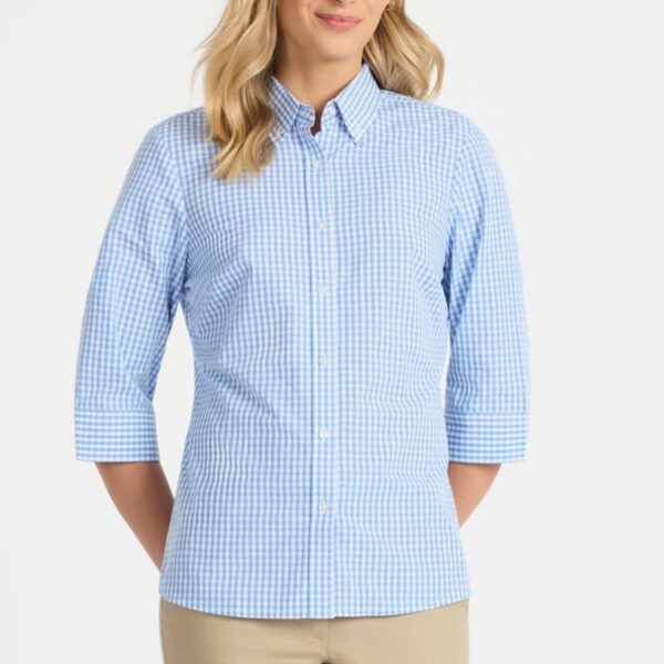 A person wearing a light blue and white checkered button-up shirt with three-quarter sleeves and beige pants stands against a plain white background, smiling with hands behind their back.