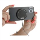 A person holding a smartphone with a wireless charger attached to the back. The charger is connected via a cable. The person has coral-colored nail polish and is wearing a silver bracelet.