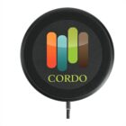 A black circular wireless charger with a multicolored logo featuring four vertical bars in shades of orange, teal, green, and brown. The word CORDO is printed in green below the logo. A charging cable is attached at the bottom.