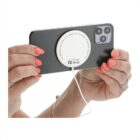 Hands with pink nail polish hold a smartphone horizontally, with a round white wireless charging pad attached to the back. A white cable extends from the pad. The phone has a dual-camera setup.
