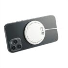 A smartphone lying face down with a wireless charging pad attached to its back. The charging pad is circular, white, and connected to a thin cable. The phone has a dark exterior and triple-lens camera setup.