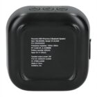 The image shows a small, square black Bluetooth speaker with control buttons on top. The back has detailed specifications including model number, battery capacity, and power output. It is made from recycled ABS material.