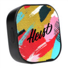 Portable speaker with a black frame and a colorful front design featuring bold abstract brushstrokes in red, blue, yellow, pink, and green. The word Heist is written across the front.