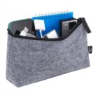Gray felt pouch with a zipper, containing a smartphone, white USB cable, small spiral notebook, silver device, and other small items. The pouch has a black tag that reads THE GOOD.